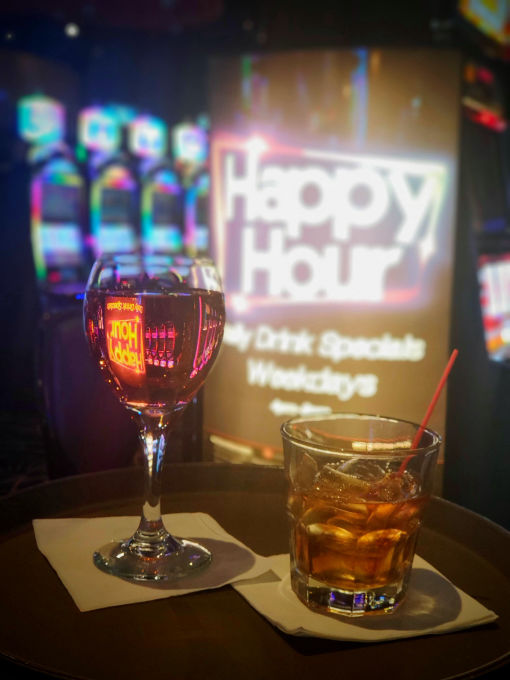 A close-up of a tray holding two drinks: a glass of wine and a mixed drink with ice and a straw. The background features vibrant slot machines and a 'Happy Hour' neon sign, emphasizing drink specials during weekdays.