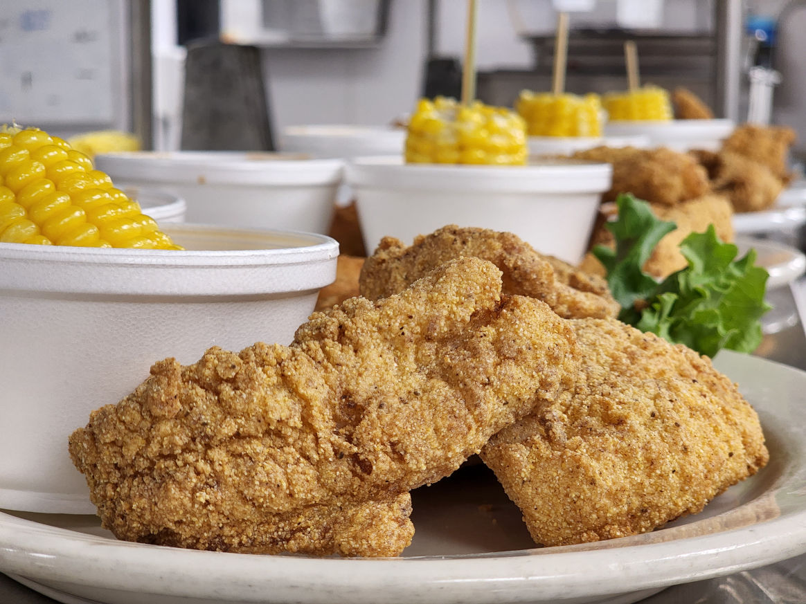A plate of golden fried catfish fillets served with corn on the cob, lettuce, and a side of sauce. The meal is presented on a white plate with a garnish of fresh greens.