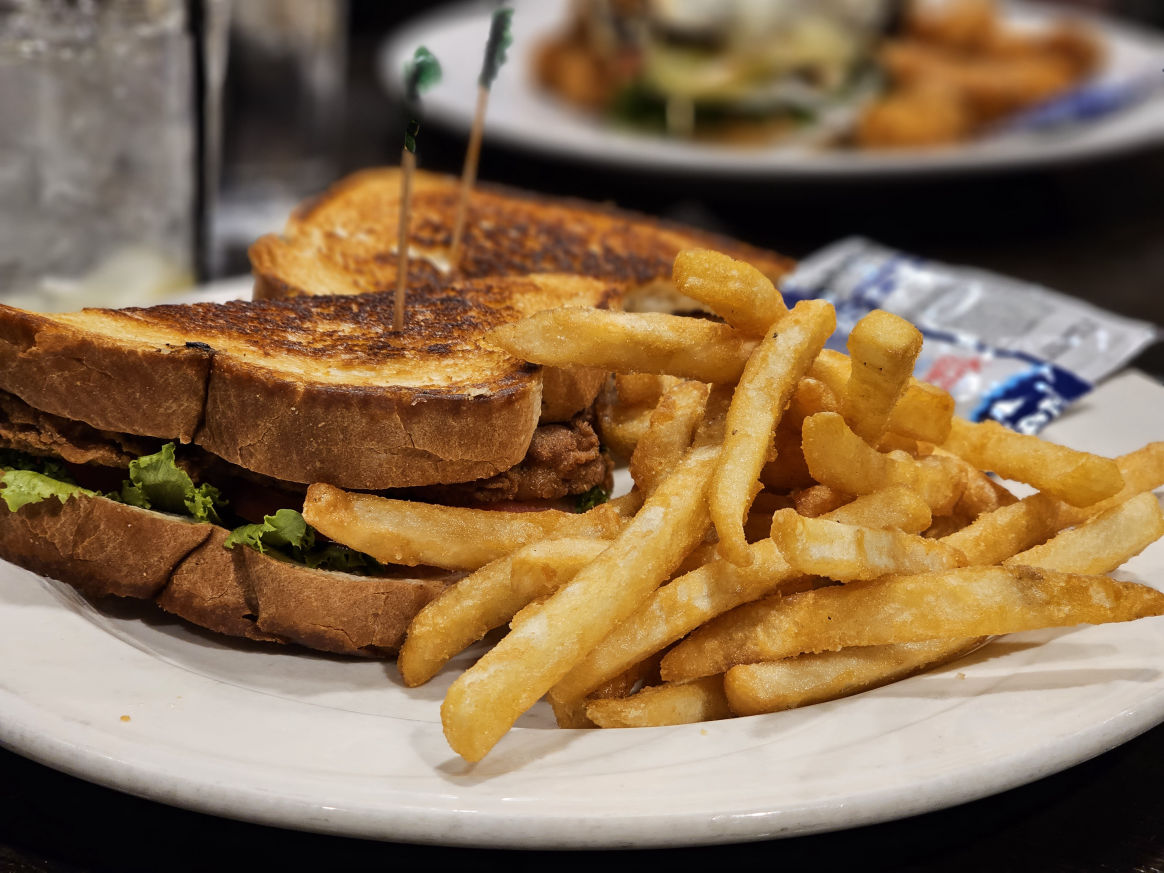 "A plate of golden crispy French fries next to a grilled sandwich with lettuce and bacon, secured with toothpicks, served on a white plate with a packet of condiments on the side."