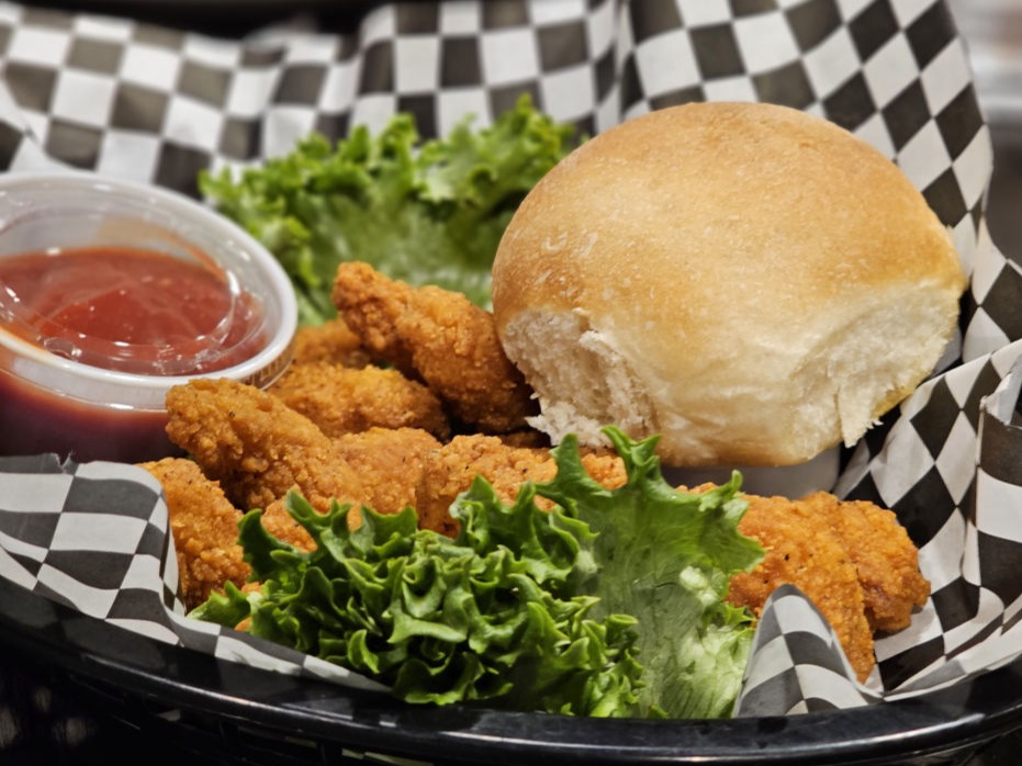 "A basket of crispy fried chicken tenders served with lettuce, a soft roll, and a side of ketchup for dipping, all wrapped in black-and-white checkered paper."