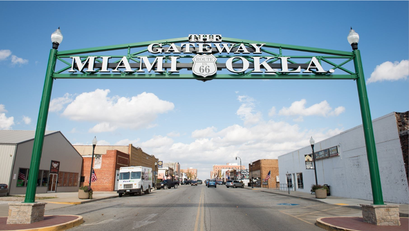 A street view of the 'Gateway to Miami, OK' arch, located on Route 66. The green arch sign prominently features the words 'THE GATEWAY MIAMI OKLA' with the Route 66 shield in the center. The scene includes a clear sky, light traffic, and buildings lining the street."