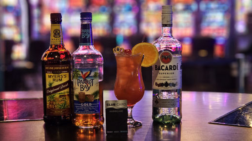 A vibrant tropical cocktail sits on the bar, made with Myers's Original Rum, Parrot Bay Gold rum, and Bacardi Superior rum. The drink is garnished with a cherry and an orange slice. In front of the drink is a High Winds Casino Premium Club Card, and the colorful background features illuminated slot machines.