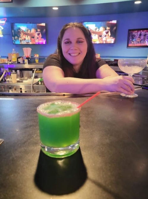 A smiling bartender behind the bar holding a margarita glass while a vibrant green drink with a straw sits in the foreground. The bar features colorful neon lighting and TVs in the background.