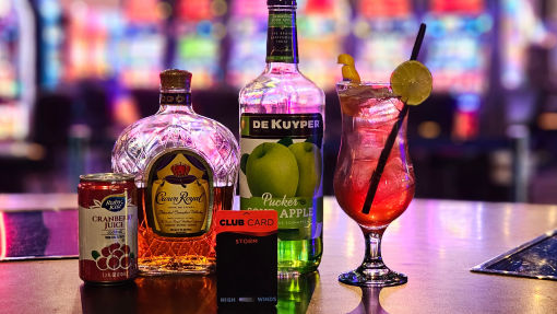 A refreshing cocktail made with cranberry juice, Crown Royal whiskey, and De Kuyper Pucker Apple liqueur sits in front of a blurred backdrop of slot machines. The drink is garnished with a lime wedge, and a High Winds Casino Storm Club Card is placed next to the bottles on the bar.